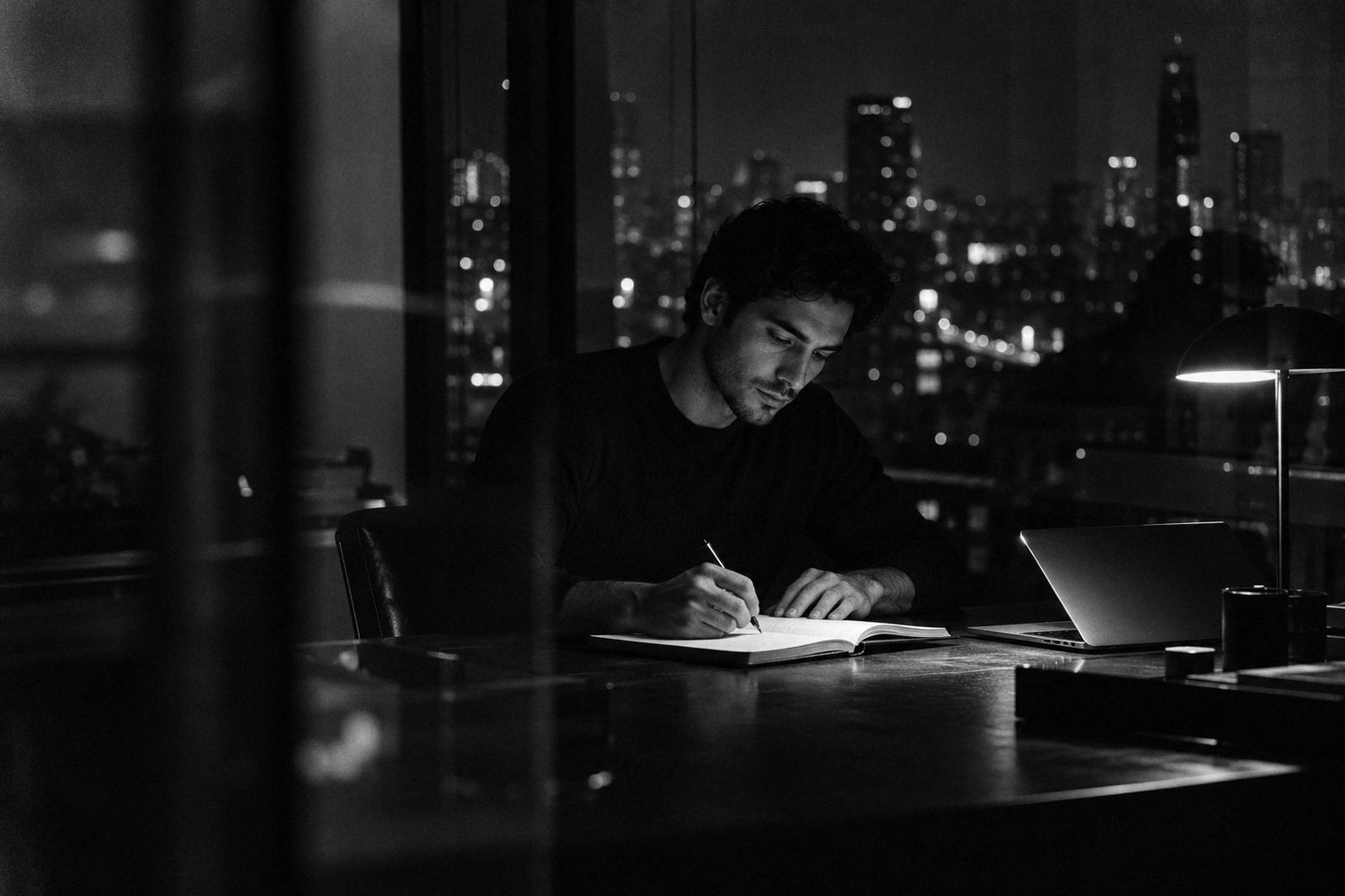 Joven estudiando de noche con lámpara y cuaderno frente a la ciudad, en blanco y negro